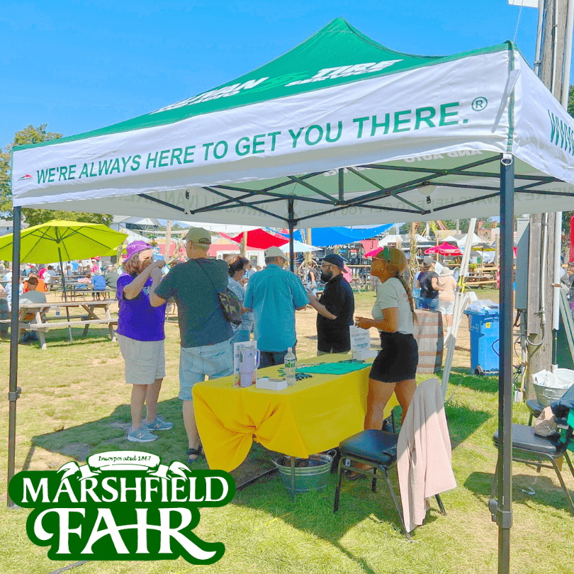 A Sullivan Tire tent set up at the marshfield fair with people underneath it talking. The marshfield fair logo in the bottom left corner.